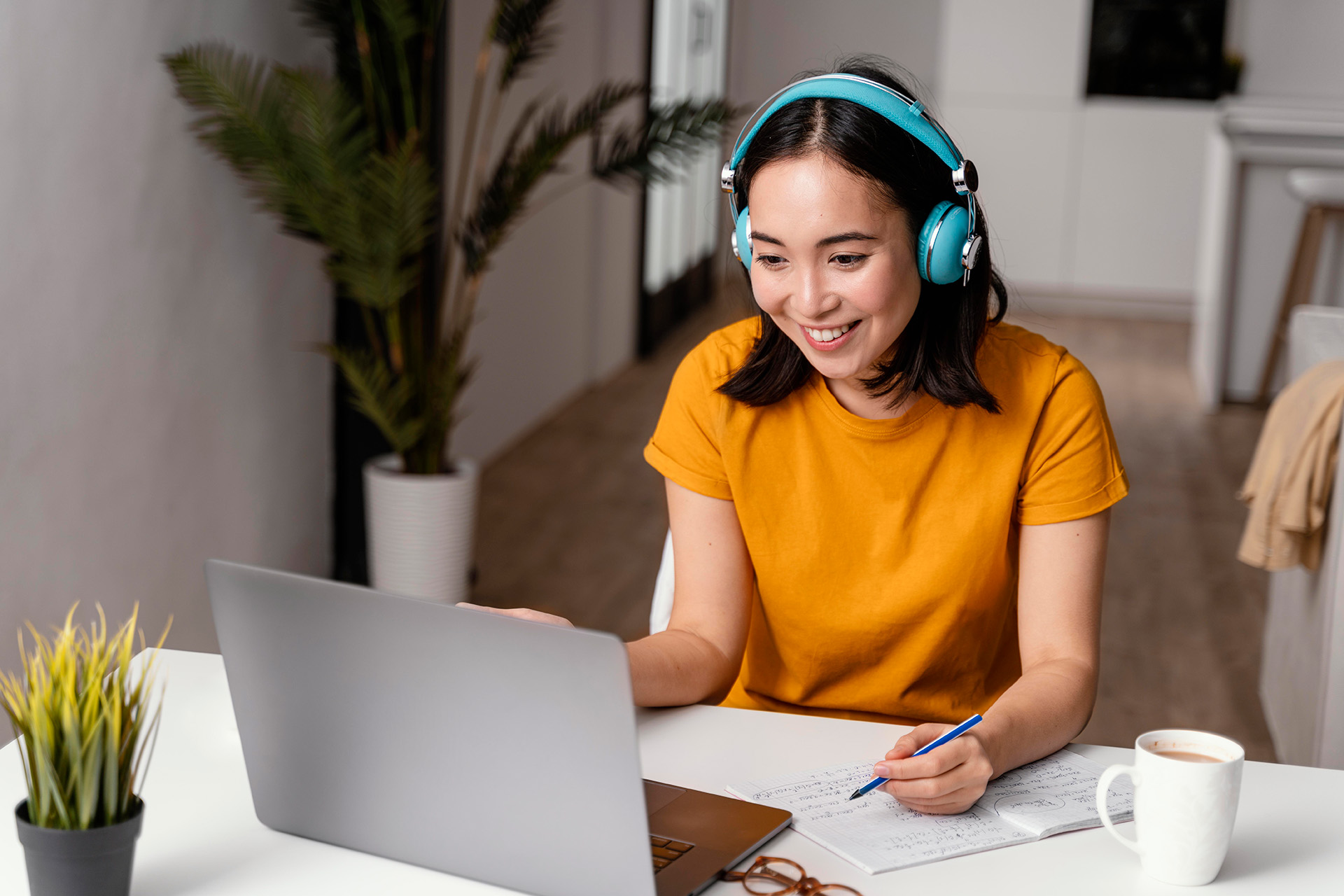 Online German courses: A young, motivated student sits at her desk at home, wearing headphones and looking at the screen of her laptop, a cup of coffee beside her.